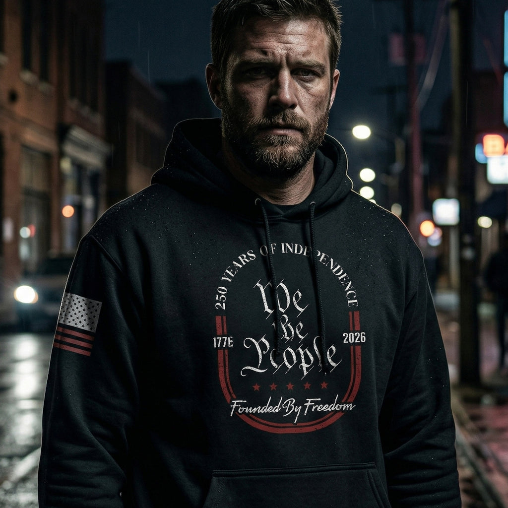 Man wearing black We The People patriotic hoodie with 250 Years of Independence design, featuring distressed typography and American flag sleeve in a nighttime urban setting